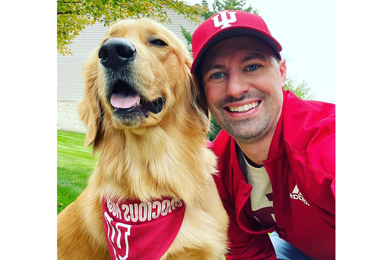 A man dressed in IU spirit wear takes a selfie with his dog, a Golden Retriever, that is wearing a red IU bandana.