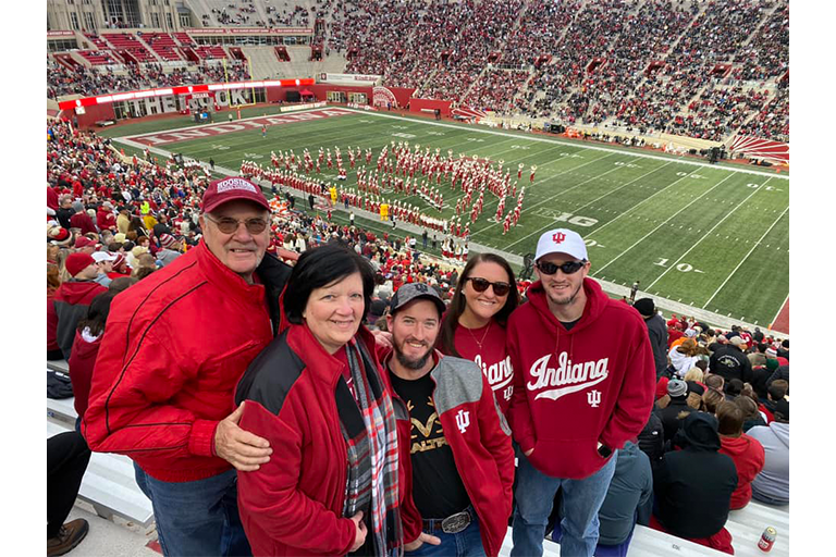 A group of five people wearing IU spirit wear smile for a photo in the stands of an IU football game at Memorial Stadium. The marching band performs on the field in the background.